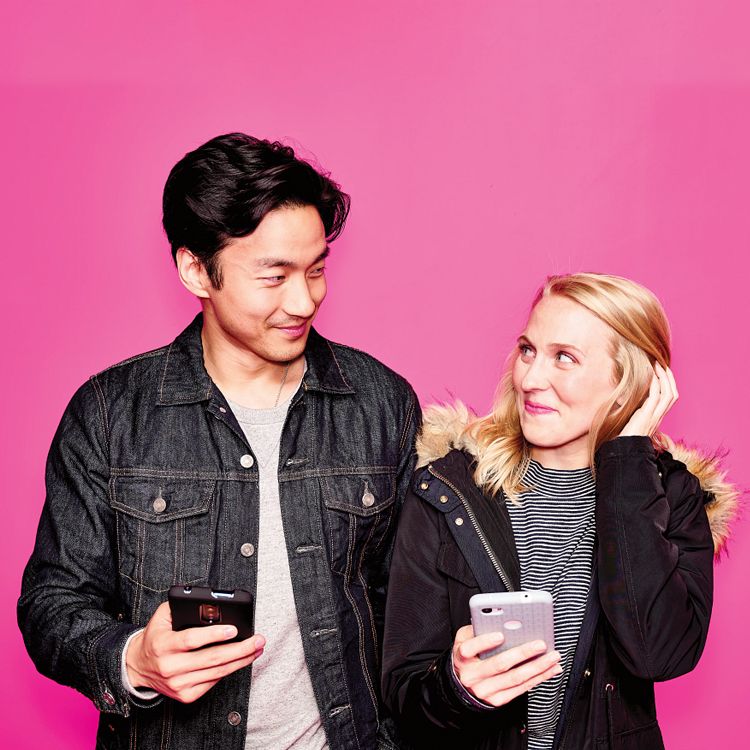 Smiling, young man and woman holding their smartphones in front of magenta backdrop.