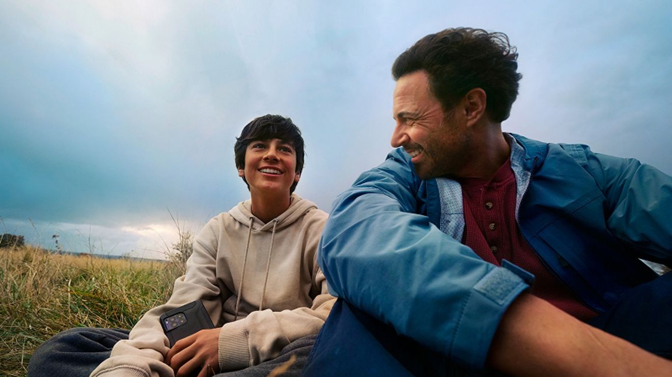 Two men hang out in a field, and one is holding a cellphone. 