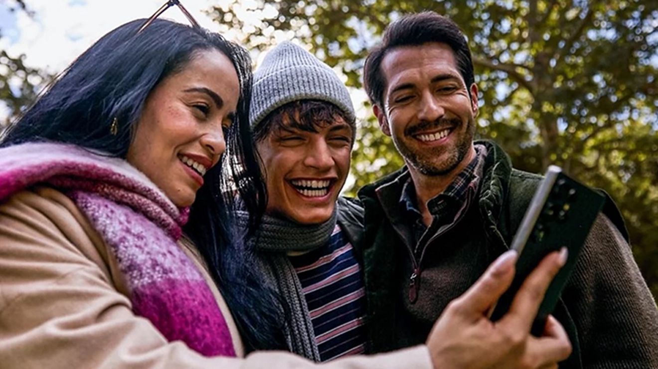 Three people look at a phone together while outside. 