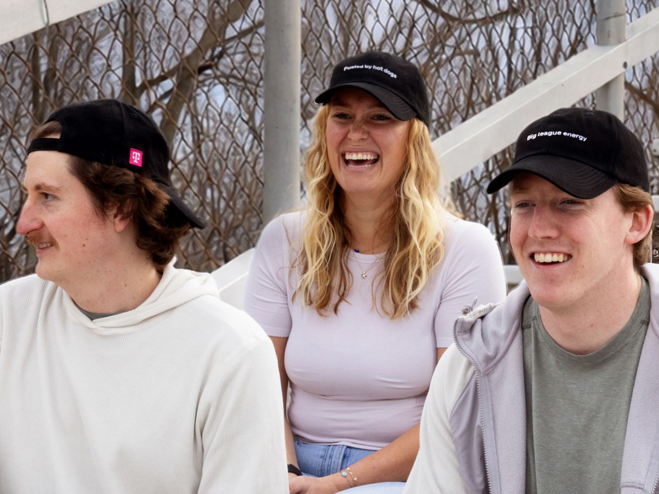 Group of 3 friends wearing Free black T-Mobile baseball hats sitting in the stands at a baseball game. Two of the hats have phrases visible on them: Big league energy and Fueled by hot dogs.