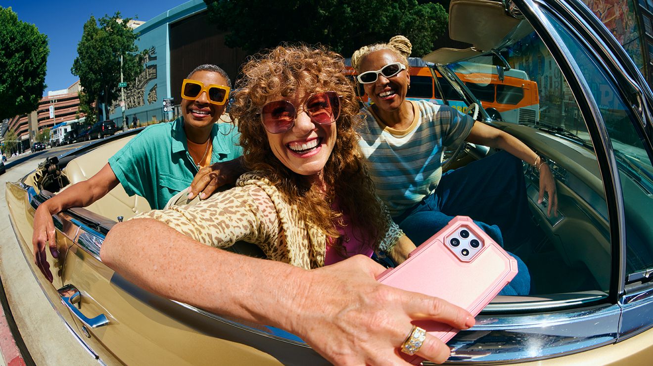 A group of three friends in an open-top convertible car. The woman in front is holding a pink smartphone.