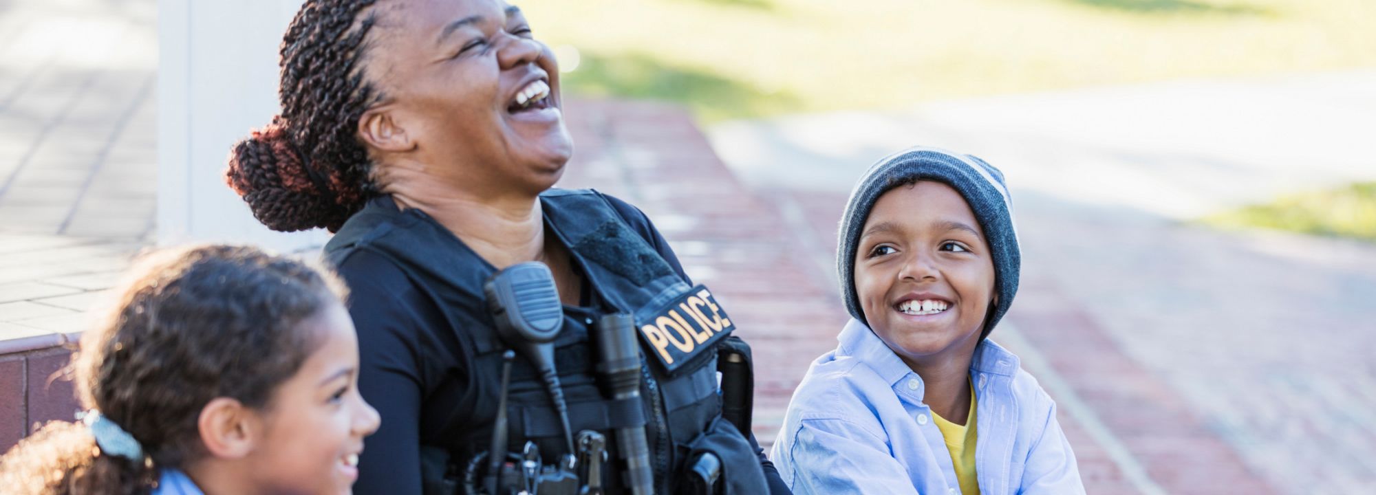 A smiling police officer sits outdoors with two children, all laughing together on a sidewalk.