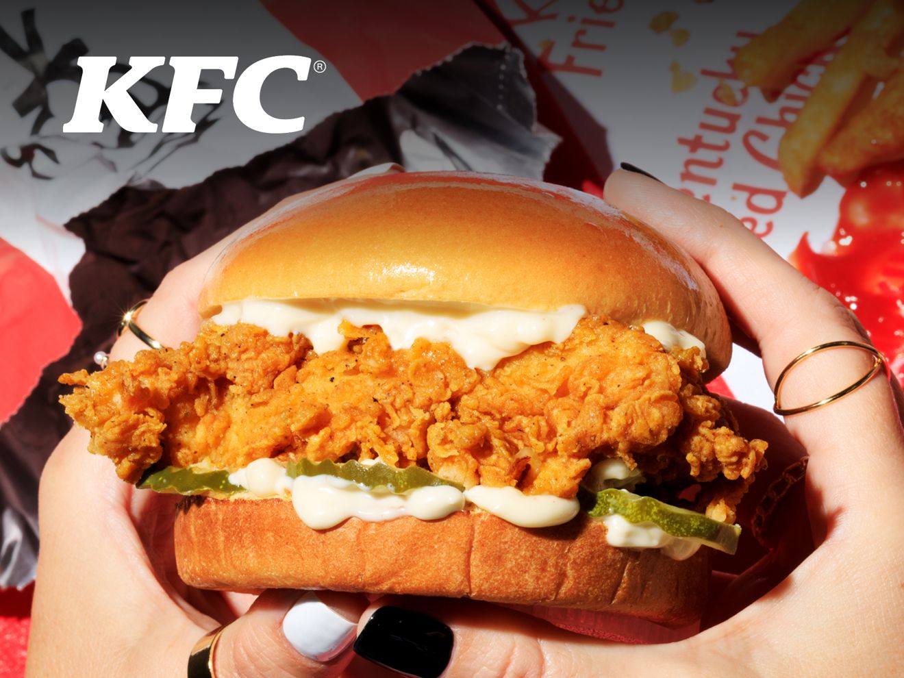 Hands holding a KFC chicken sandwich above KFC wrappers and fries