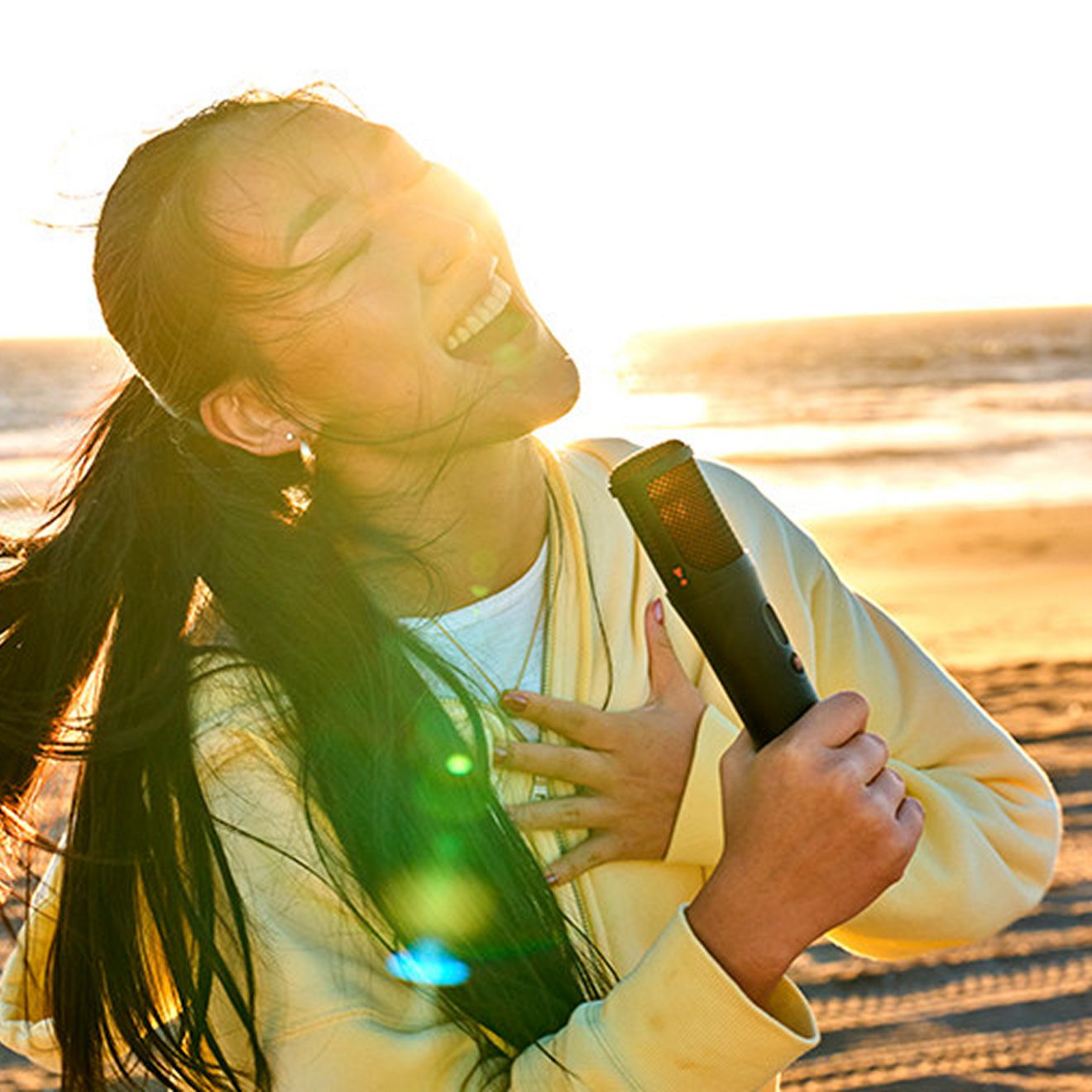 A woman sings on the beach with the sunset behind her using the JBL PartyBox On-the-Go 2.