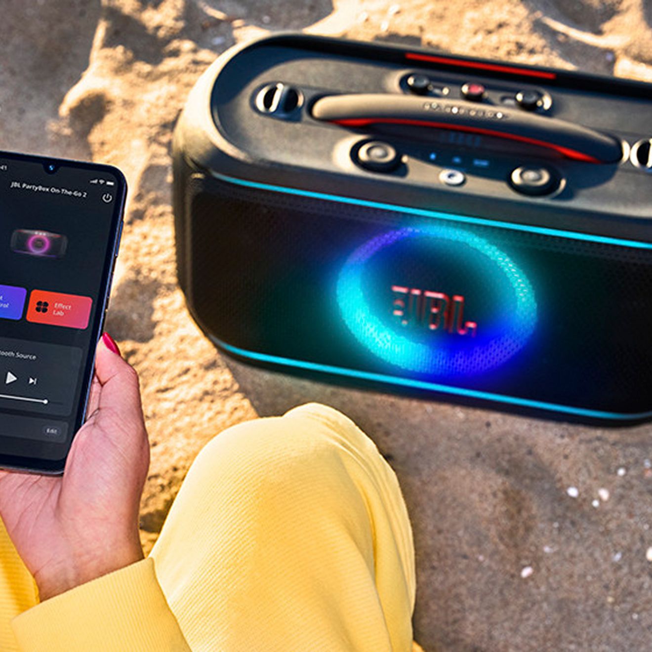 A woman is on the beach and uses the JBL PartyBox App on her phone to play music on the JBL Partybox On-the-Go 2.