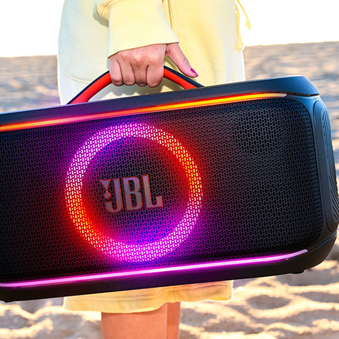 A hand carries a JBL PartyBox On-the-Go 2 while walking on the beach to their destination.