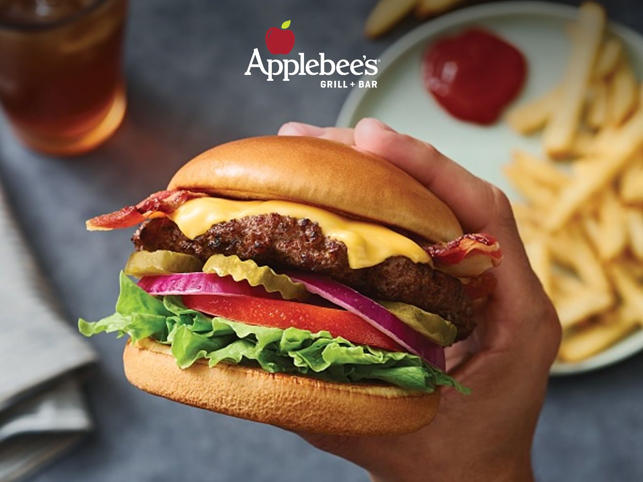 Closeup of a hand holding an Applebee’s Classic Burger with a plate full of fries on a table in the background. Applebee’s Grill and Bar logo hovers above the burger.