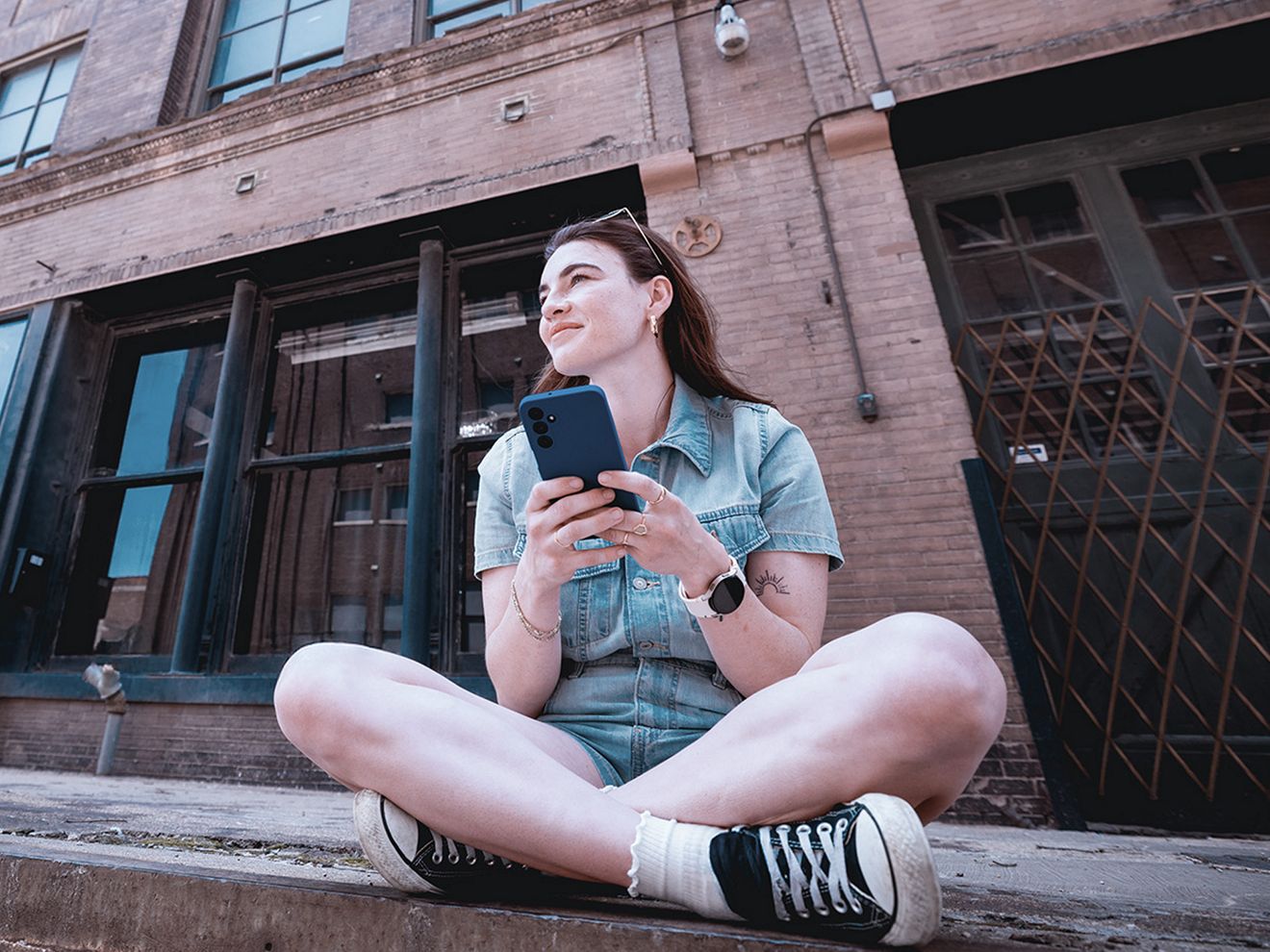 A woman sits cross-legged outside on a city street corner while enjoying using her smartphone.
