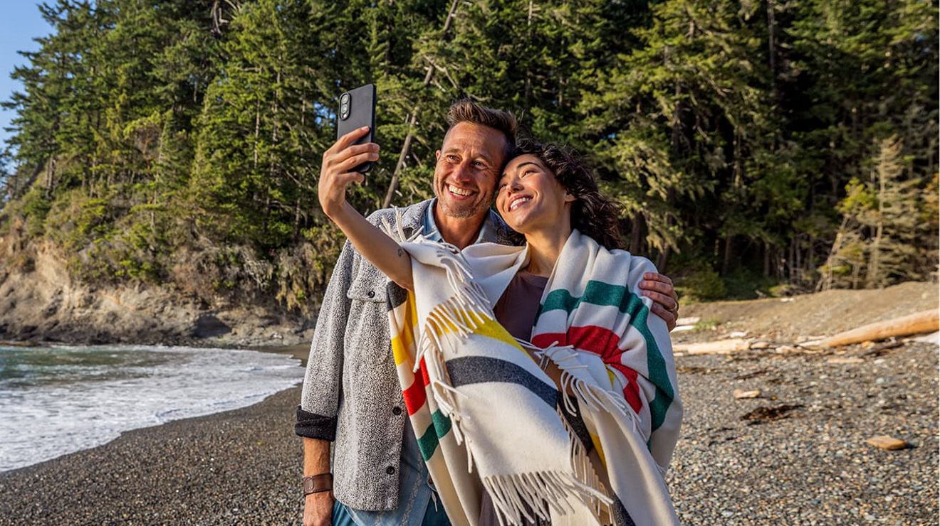 Under a beautiful blue sky, a woman and a man pose for a selfie on a pebble beach.