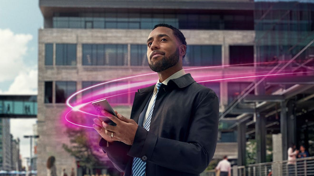A professionally dressed man standing outside a modern office building looks at his smartphone while a glowing pink light trail swirls around him, suggesting digital connectivity or wireless technology.