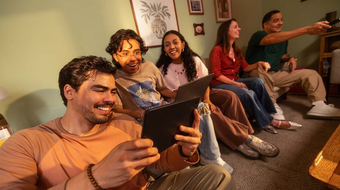 Group of friends gathered in a living room, using smartphones and watching a screen while sitting together on a couch and floor.