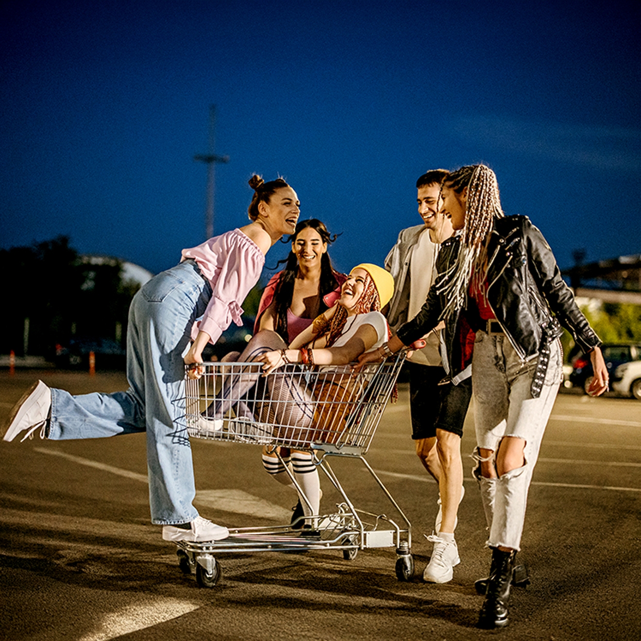 A group of friends having fun at dusk, pushing a shopping cart across an empty parking lot with big playful energy.