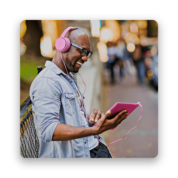 Smiling man holding magenta tablet while listening to magenta headphones on the sidewalk