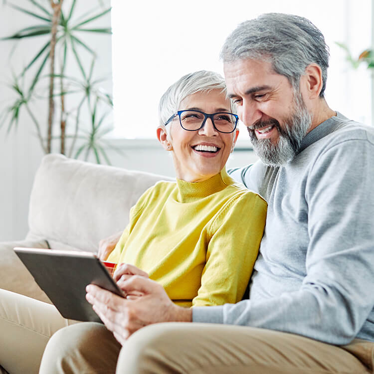 On the couch, a senior couple smiles while looking at a laptop.