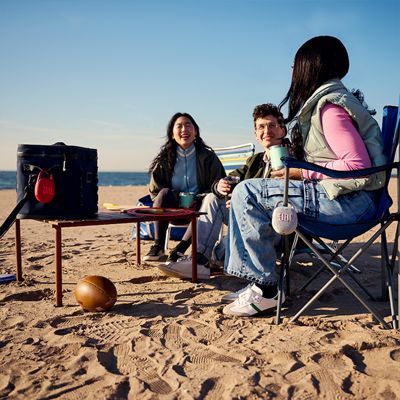 Two women and a man are at the beach listening to music with a JBL Clip five.