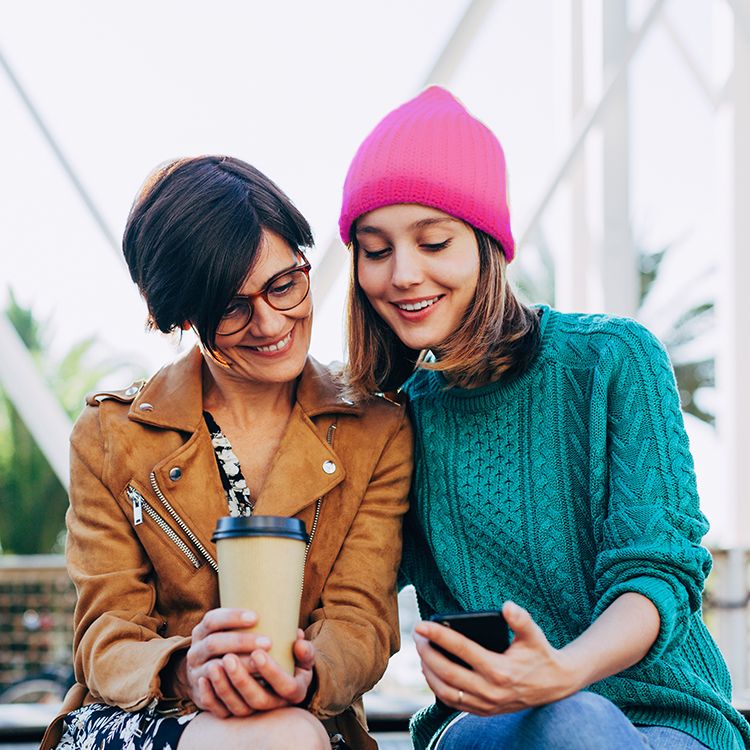 Two women hold coffee and smile while looking at a phone.