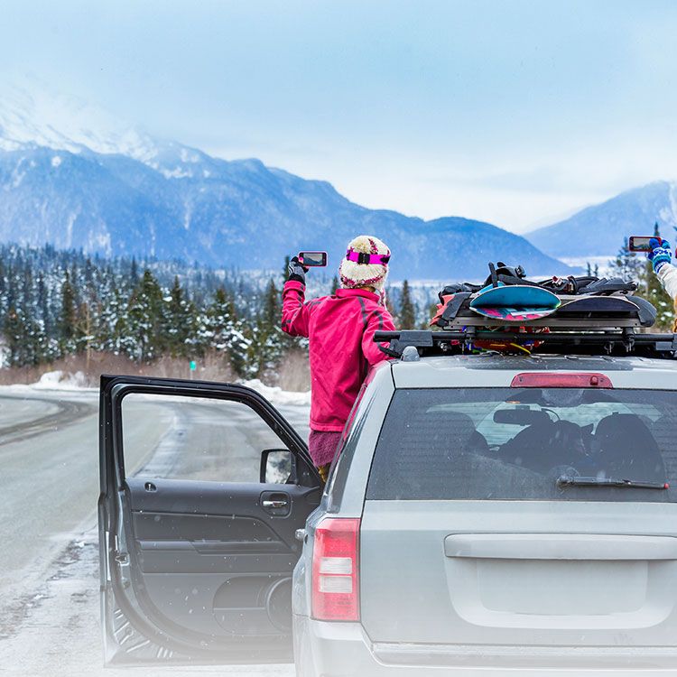 Car on the road in a snowy landscape with mountains; woman leaning out of car taking picture of mountains with phone.