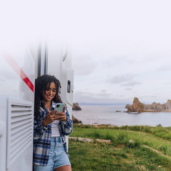 Young woman checking her phone with camper and ocean in the background.