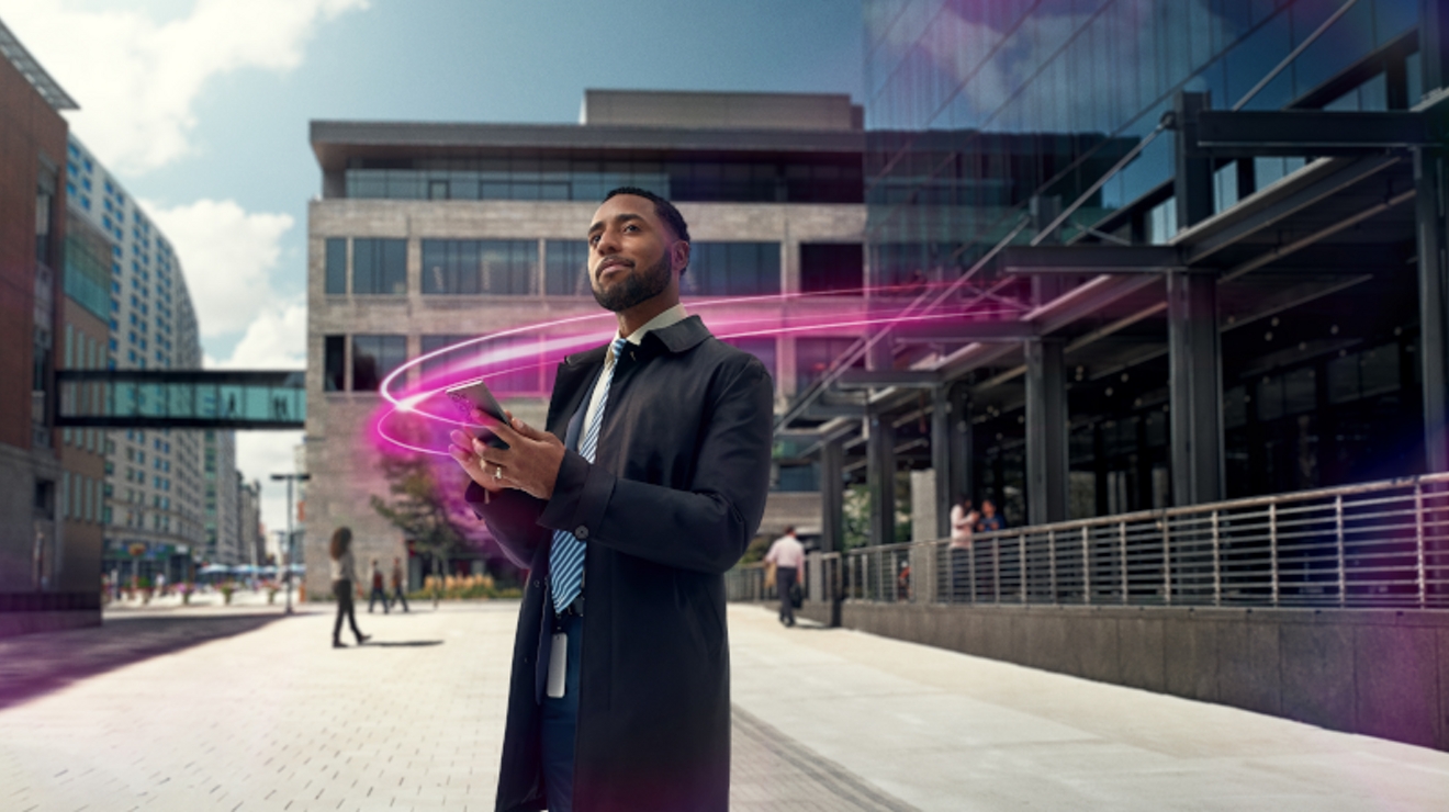 A man in a black overcoat holding a cellphone outside an office building with magenta flashes in the background.