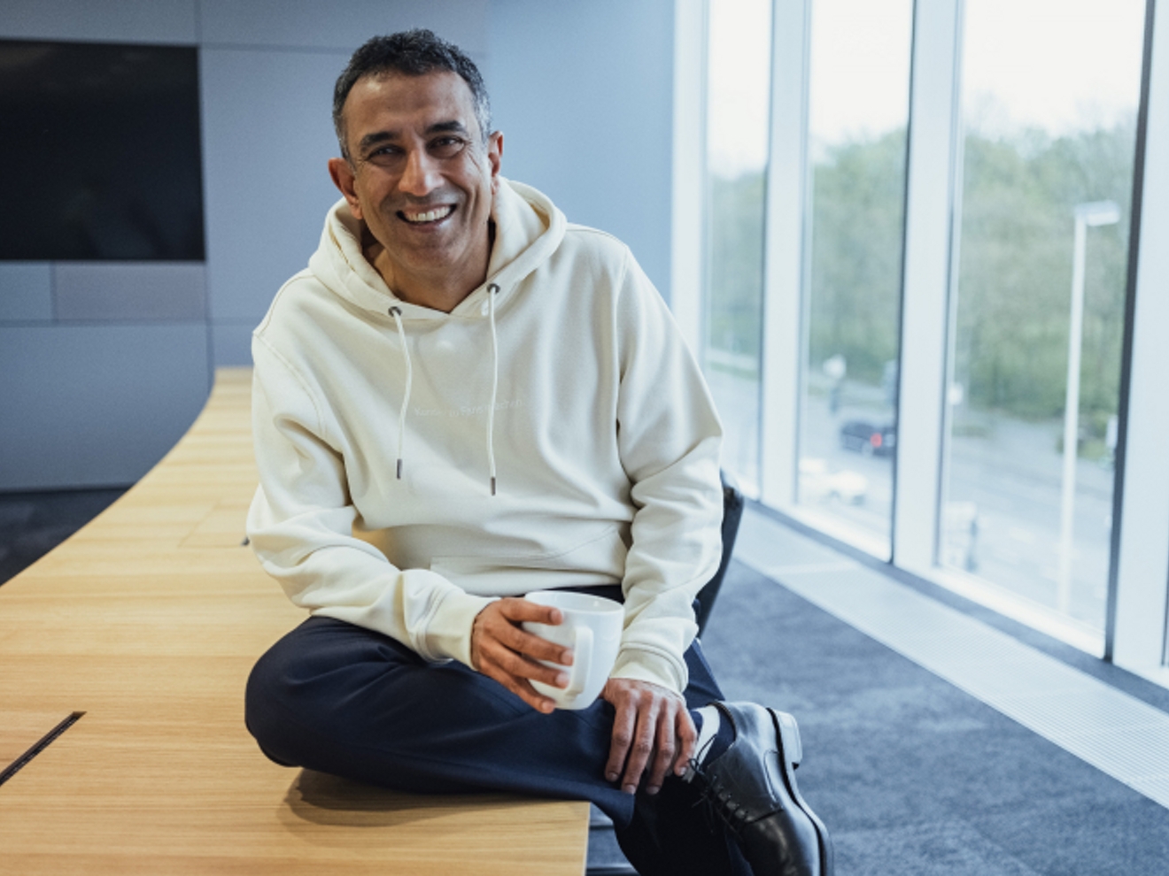 Srini smiling while holding a coffee mug and leaning casually against a table.
