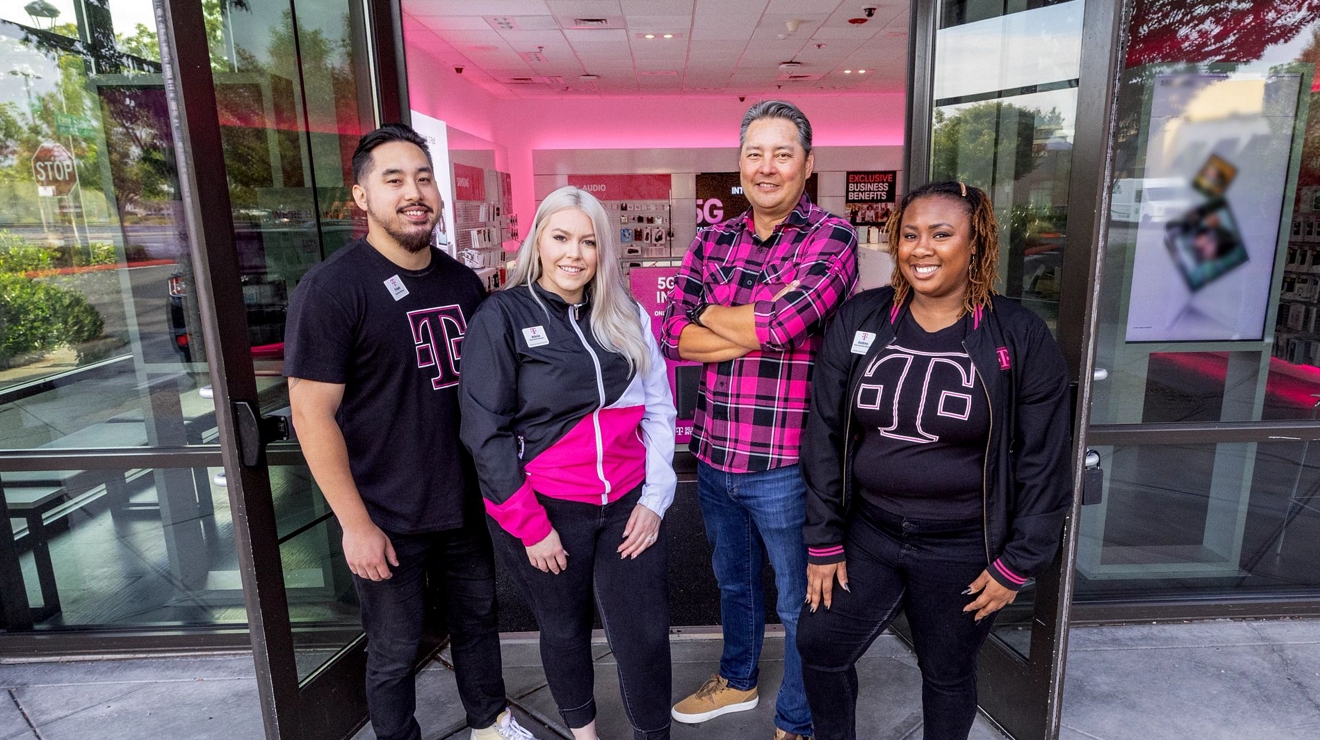 A group of T-Mobile employees are standing together in front of a T-Mobile store, smiling.