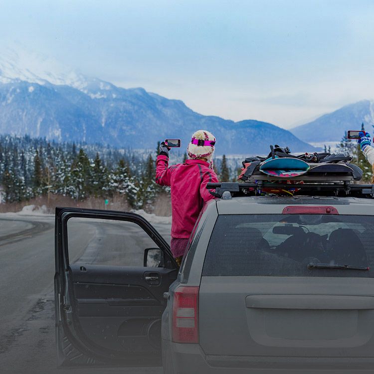 Mujer en una ruta nevada, inclinándose fuera de un SUV para tomar la foto de una montaña con el teléfono.