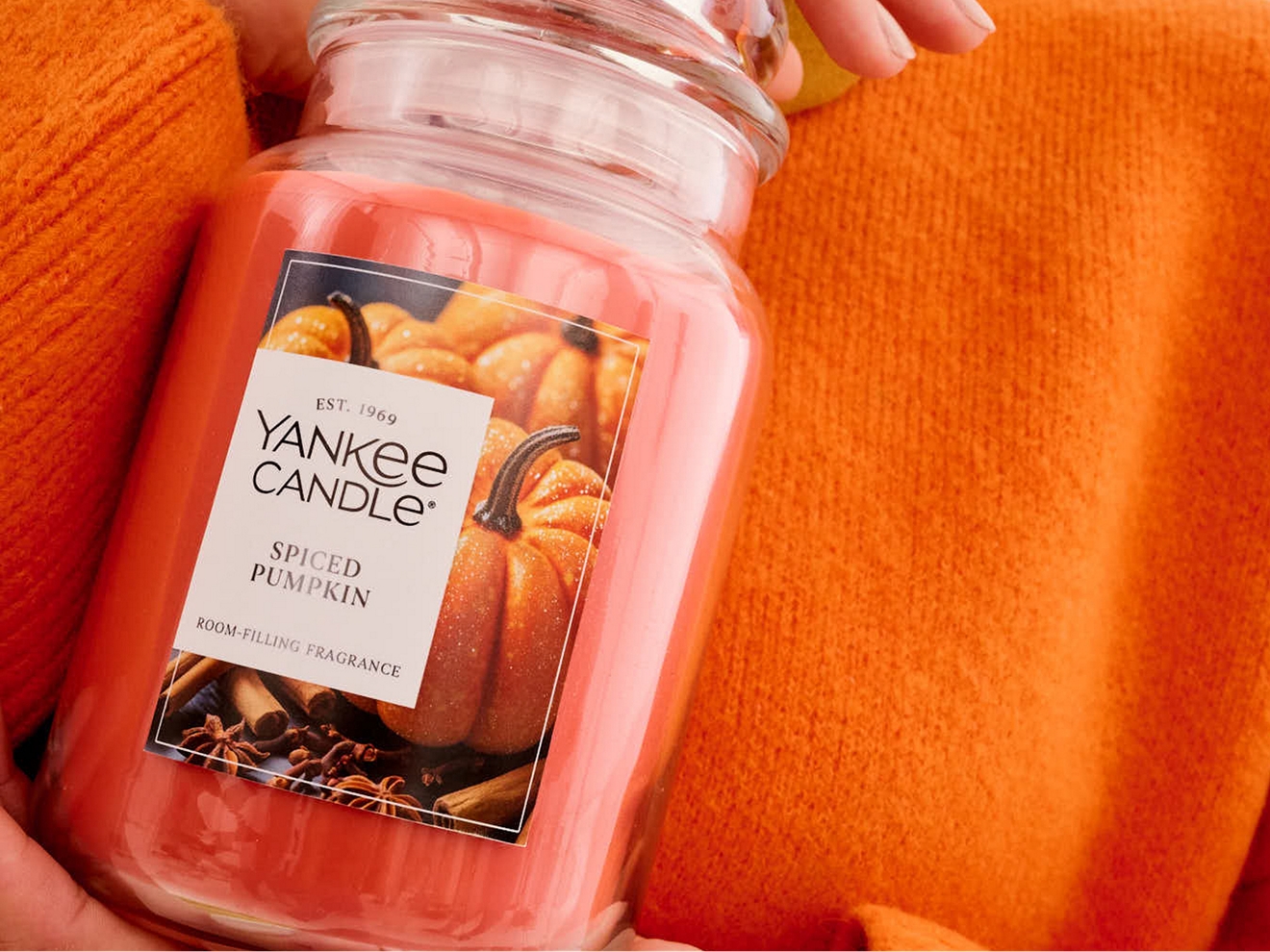 A pair of hands holding a large orange pumpkin spice glass candle.