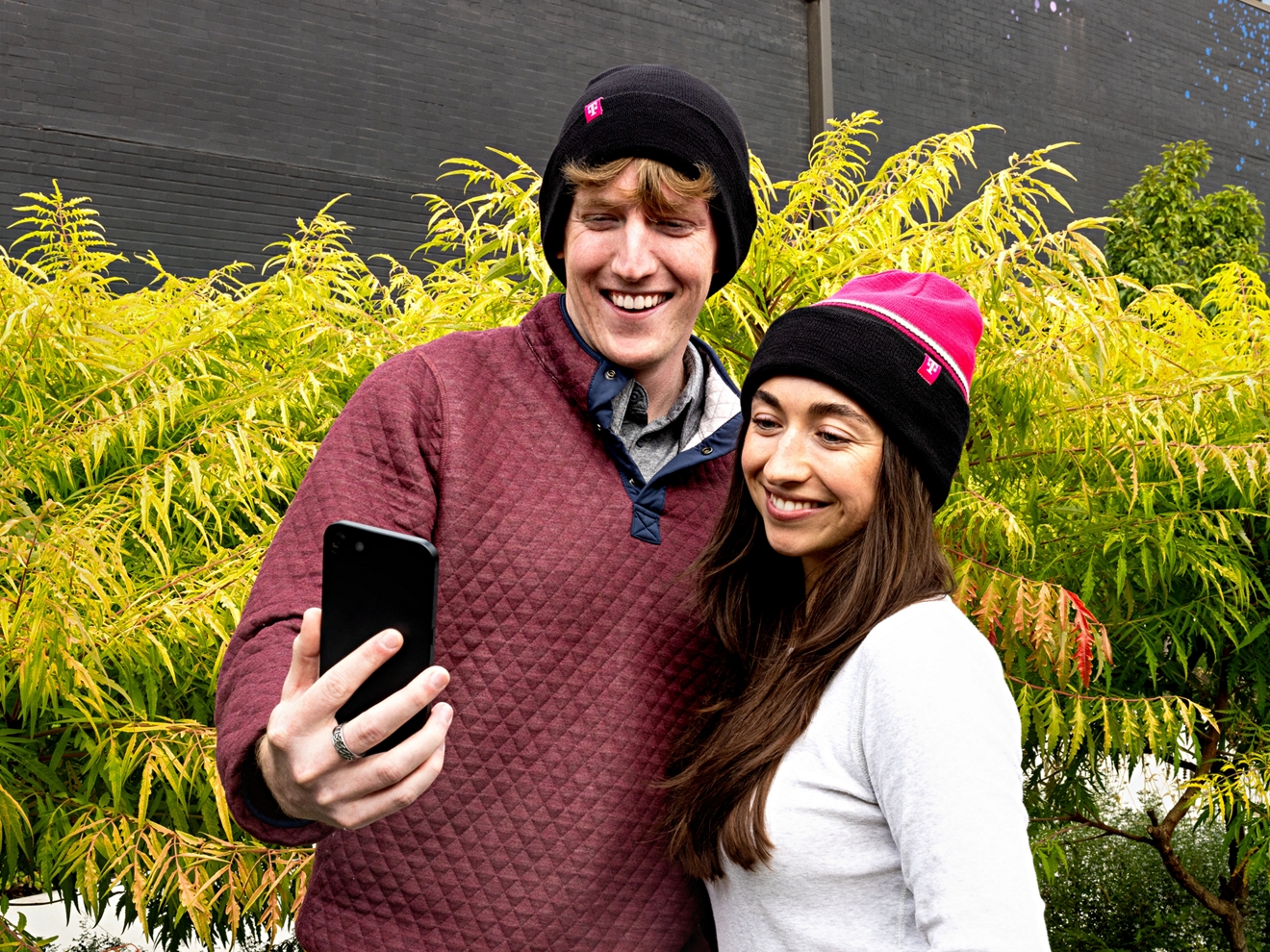 A couple wearing reversible beanies poses for a selfie outdoors.
