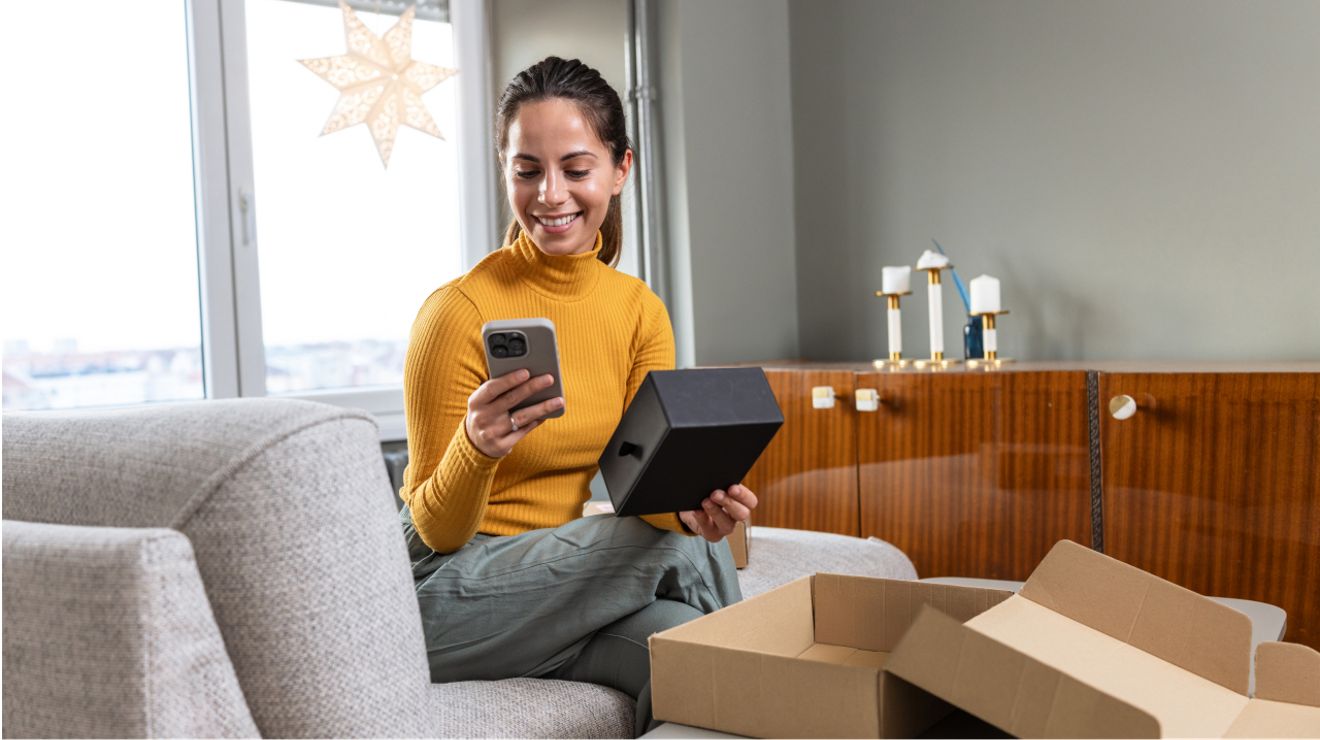 A woman in an orange turtleneck smiles down at a new phone she’s just unboxed as she sits in her living room.