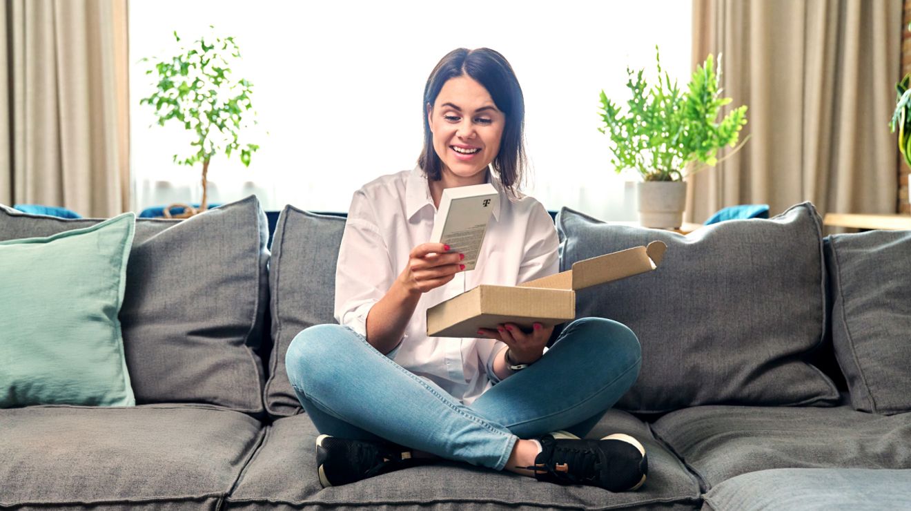 A smiling woman in a button-down shirt and jeans sits cross-legged on her couch as she unboxes a new phone.