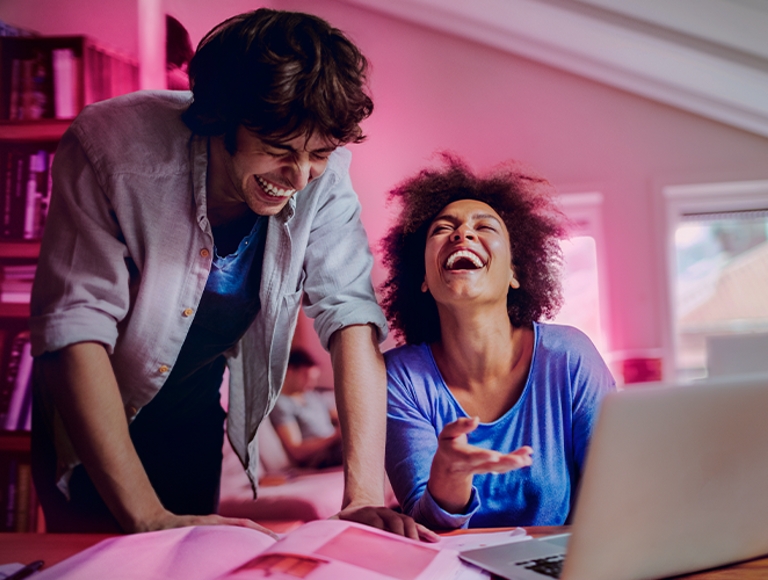 ​​Two college students laughing while doing schoolwork on a laptop at home.​