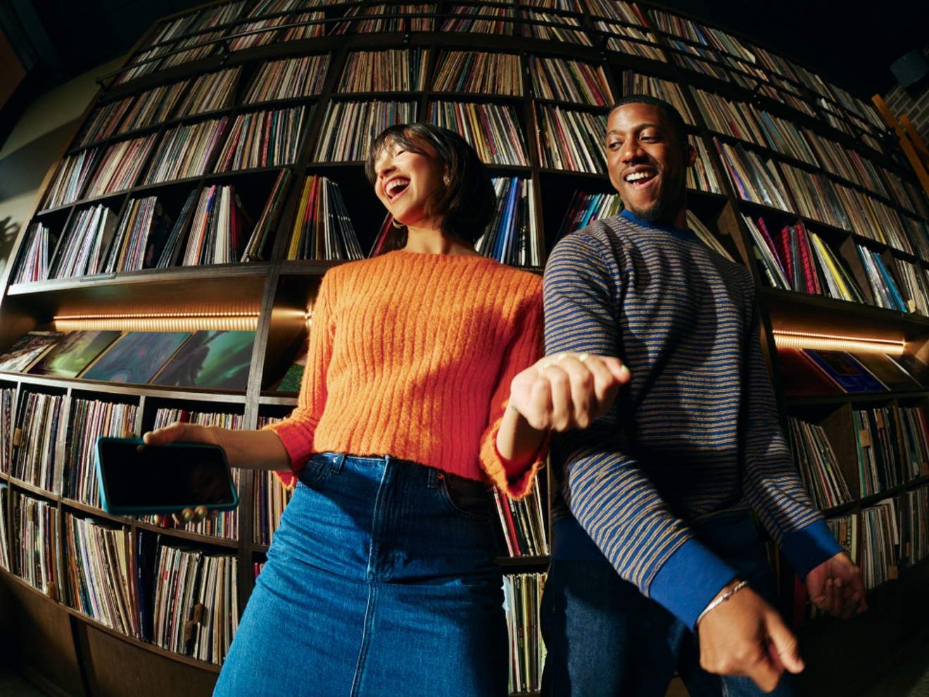 A woman in an orange sweater and man in a striped sweater smile and dance in front of a wall of vinyl records on shelves. She holds a blue phone in her hand.