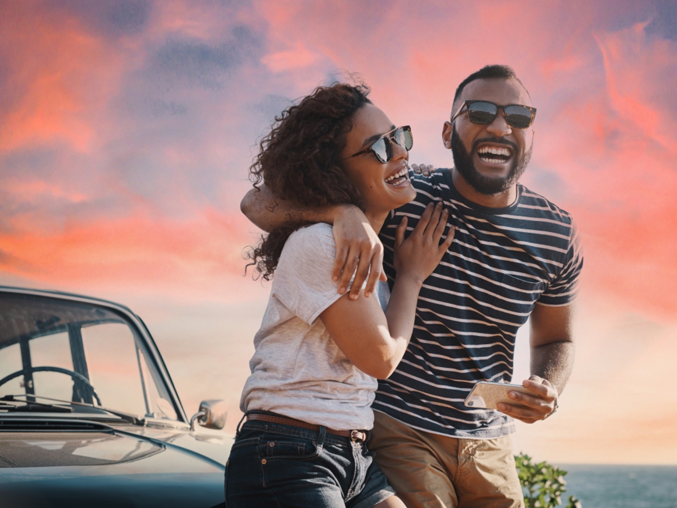 A couple are laughing while sitting on their parked car. It is evening with a sunset behind them.