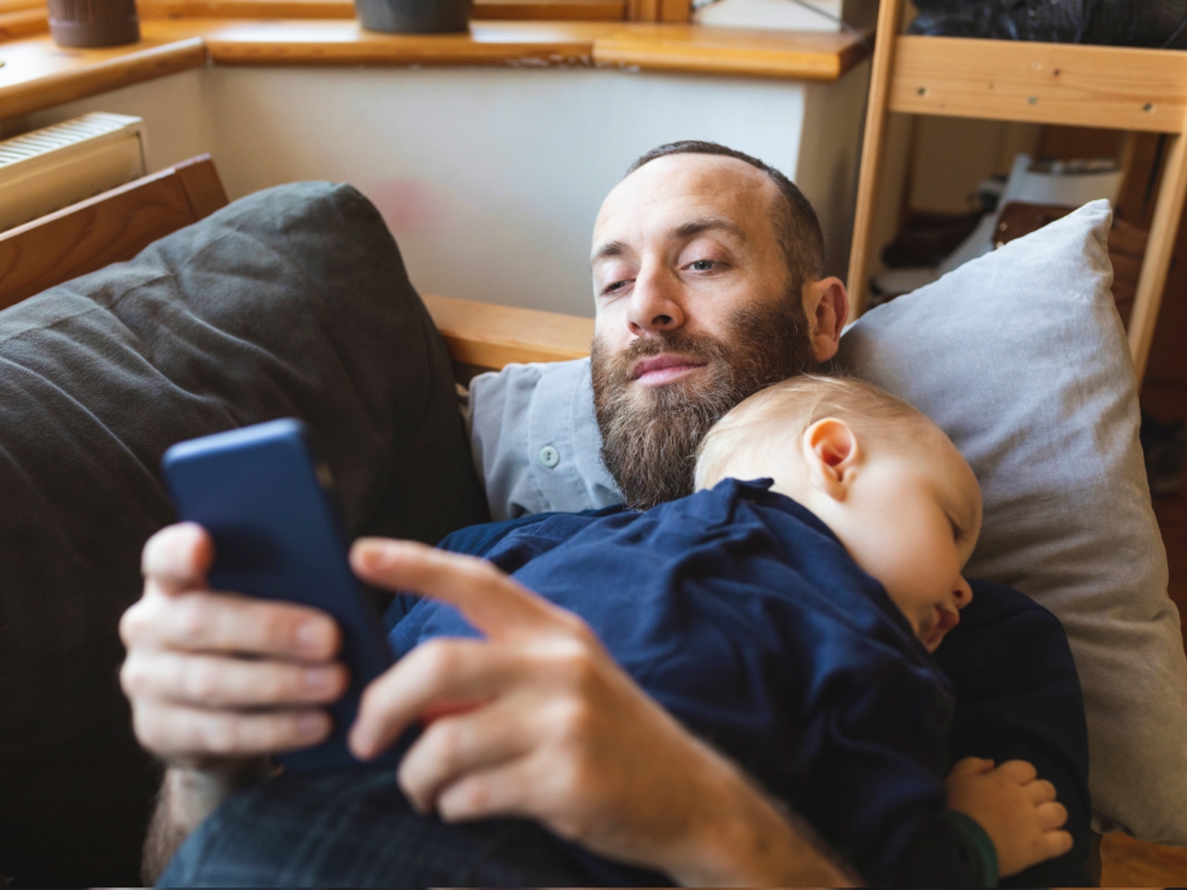 A father is laying on a couch with a baby sleeping on top of him. He is on his cell phone.