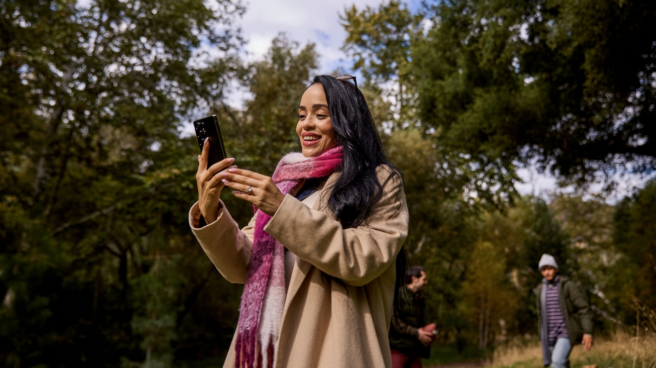A woman in a magenta and white plaid scarf stands outside, surrounded by trees, smiling at her phone.