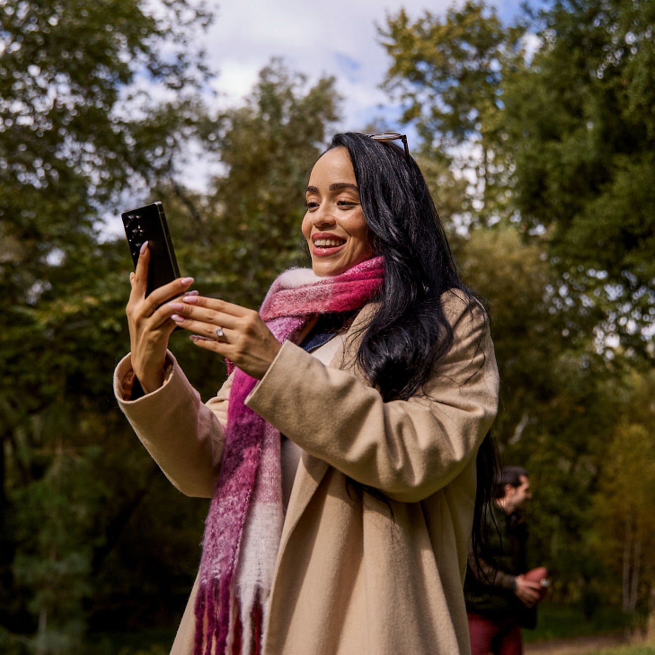 A woman in a magenta and white plaid scarf stands outside, surrounded by trees, smiling at her phone.