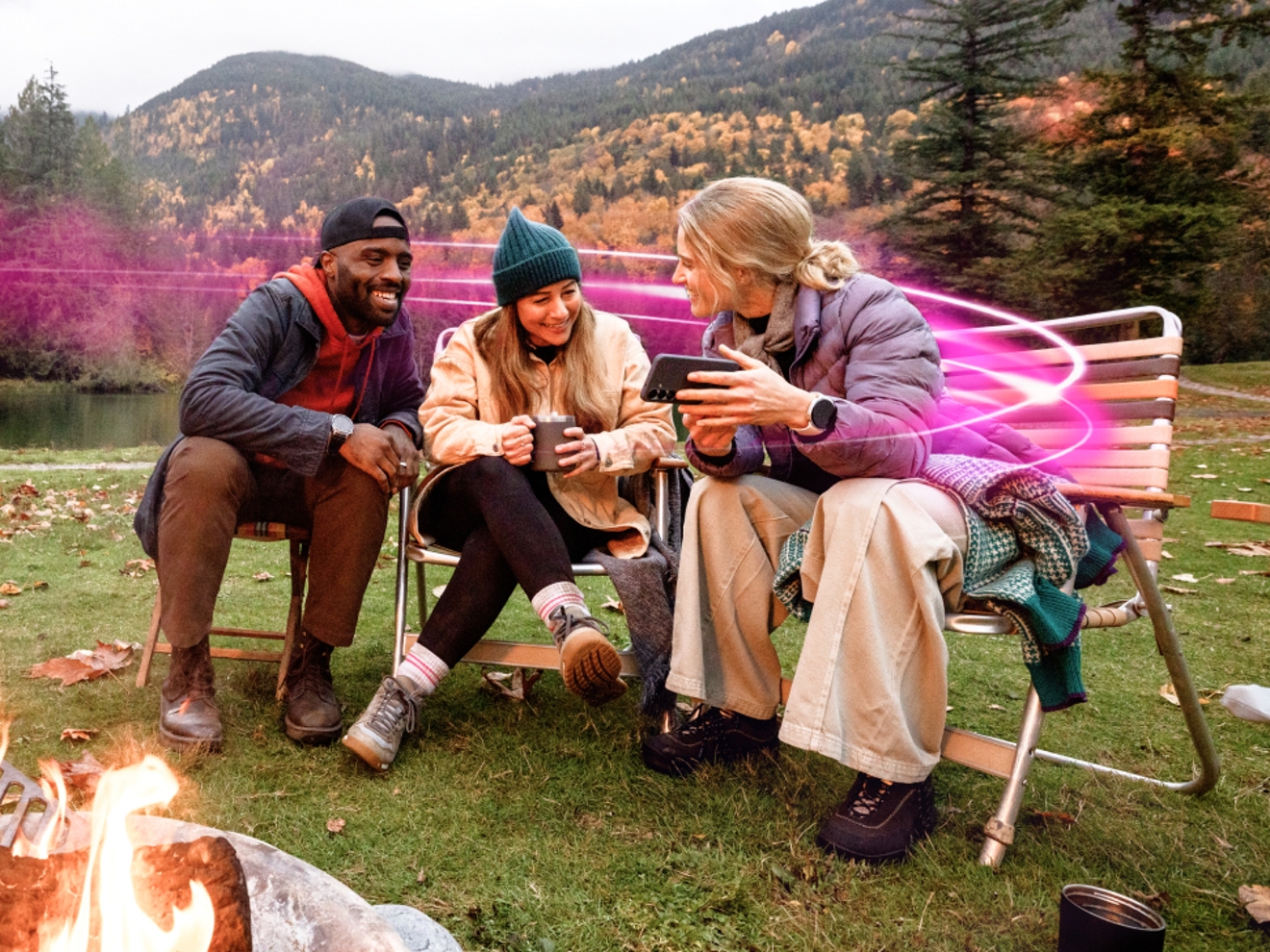 Three friends relaxing by a campfire in the mountains, bundled up in warm layers as they lean in to check something on a phone, with a soft magenta glow curving behind them.