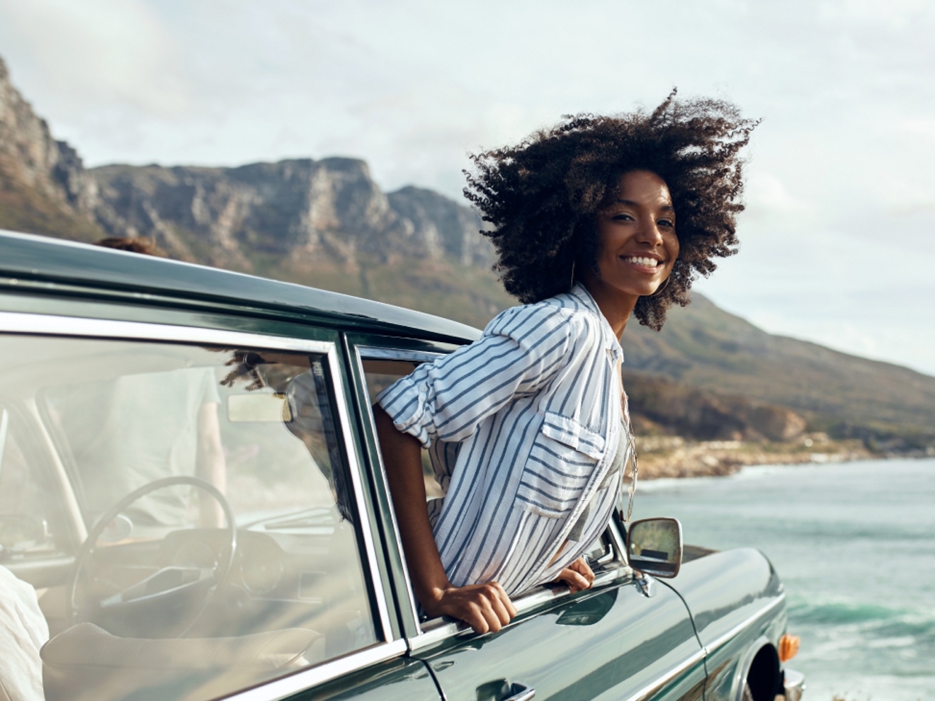 A smiling woman sticking out of a parked car window while at the beach.