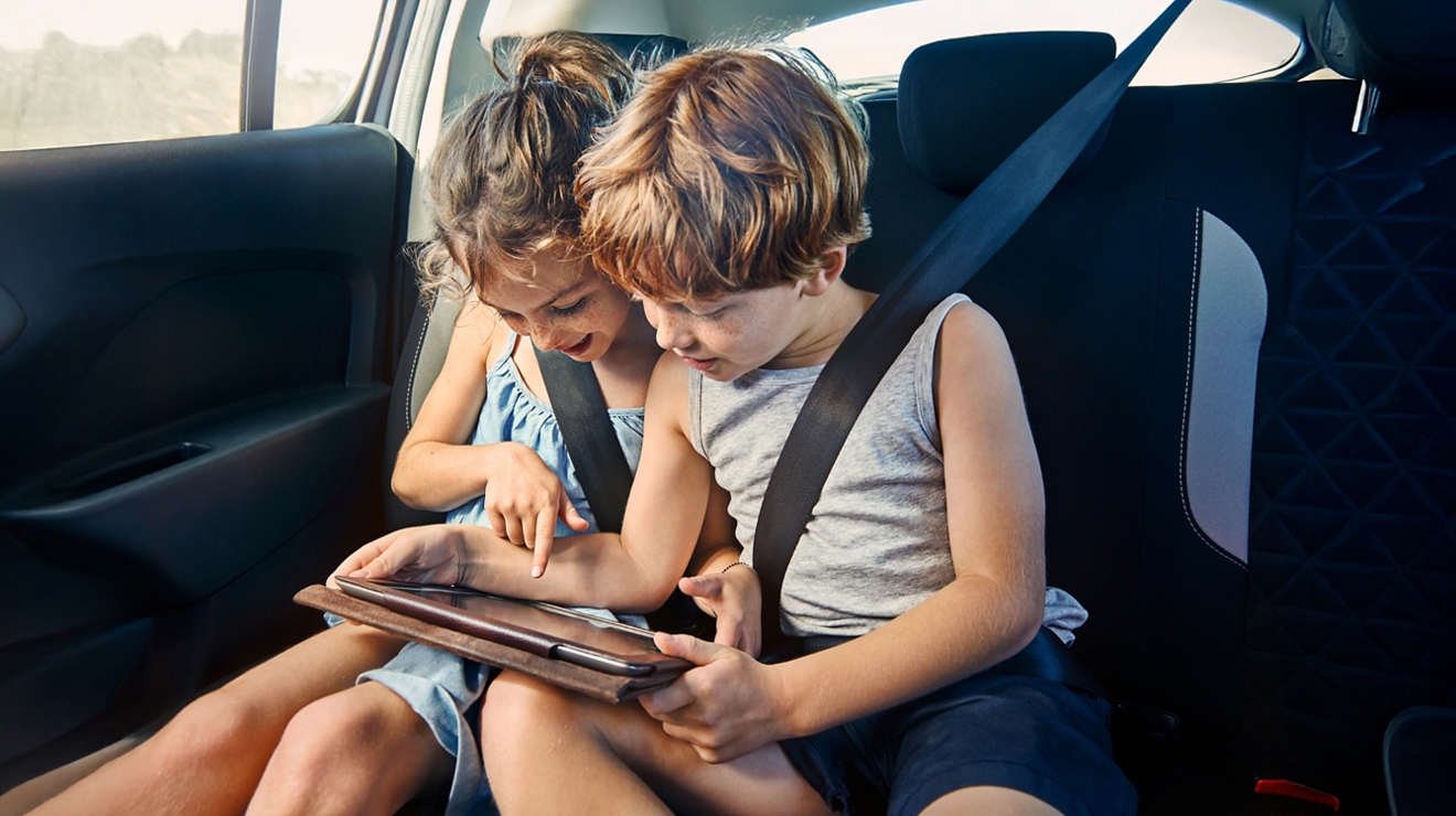 Two kids in the backseat of a car, watching a tablet while interacting with the screen.