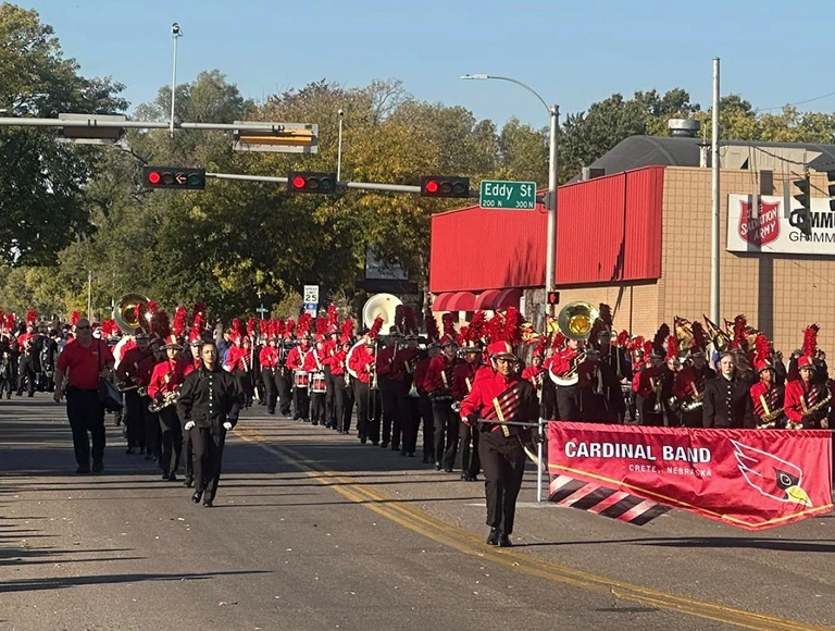 The Crete High School marching band, wearing red and black, marches down the street during a parade.