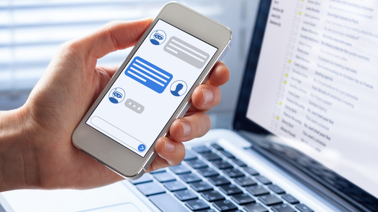 A person’s hand is holding onto a smartphone that displays a chat conversation with message icons. A laptop with informational content is displayed on the screen behind the person’s hand.