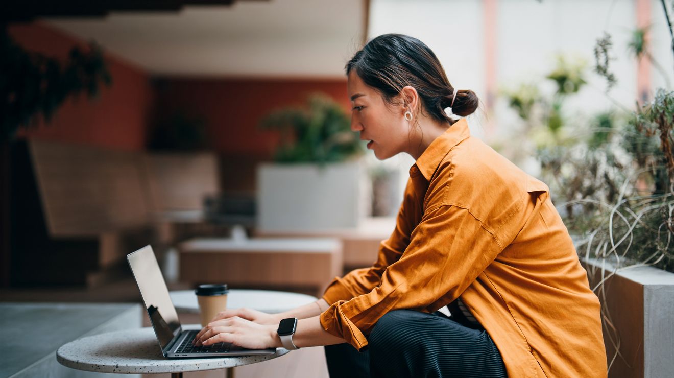 Woman working on a laptop at a small round table with a coffee cup beside her in a casual indoor  setting