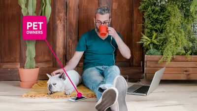 Man sitting on the ground, drinking coffee and petting the dog lying next to him. A magenta text bubble connected to the dog and man reads “Pet owner.”