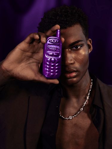 A dark-skinned man holds up a purple fragrance bottle shaped like an early 2000s-era cellphone over his face, with his shirt half un-buttoned.