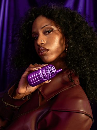 A dark-skinned woman holds up a purple fragrance bottle shaped like an early 2000s-era cellphone with a purple velvet background.