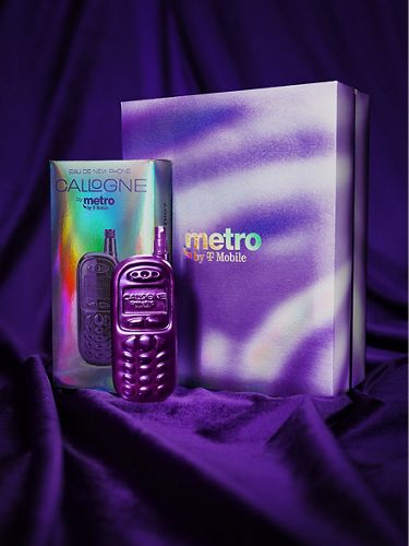 A purple fragrance bottle shaped like an early 2000s-era cellphone sits in front of iridescent Metro by T-Mobile boxes, with a purple velvet backdrop.