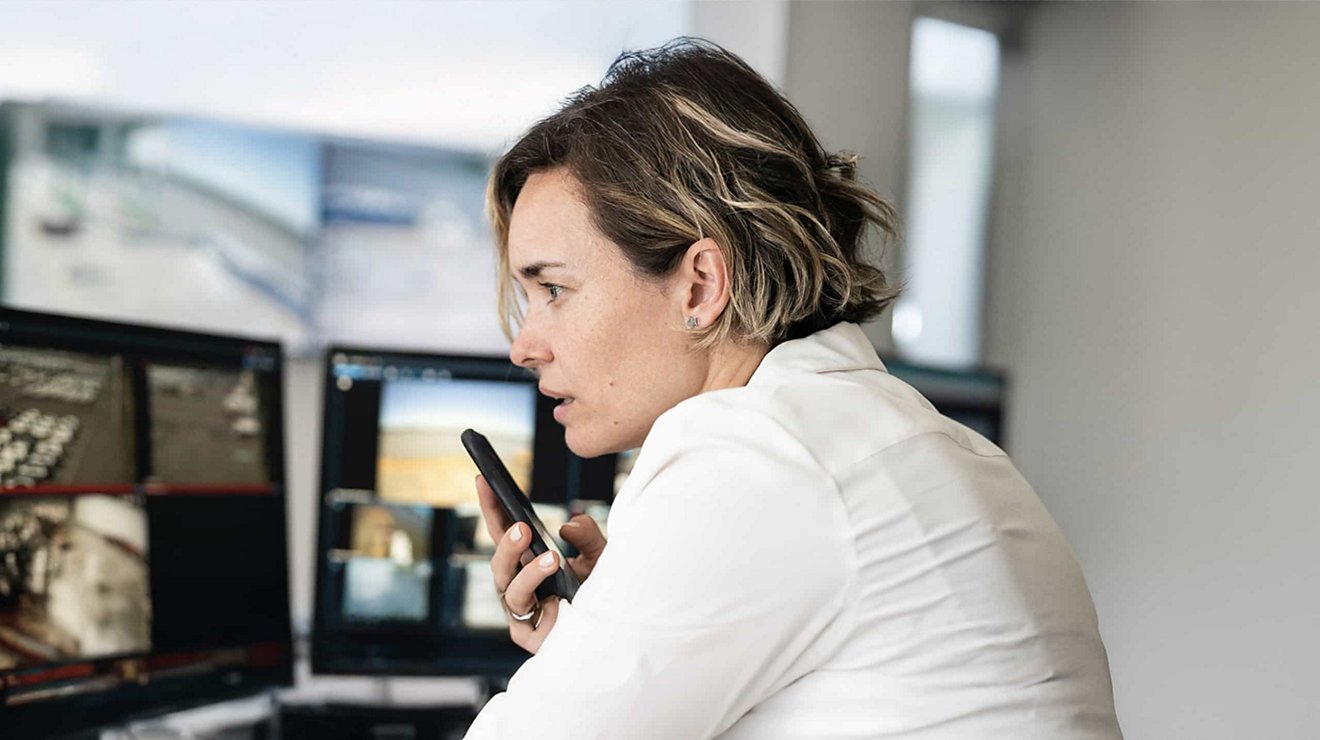 A professional uses speakerphone on a mobile device from a workstation with multiple monitors.