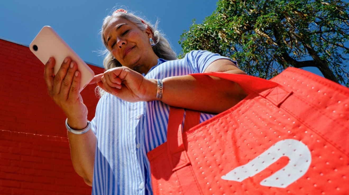 A woman in a blue and white striped shirt smiles at her phone as she holds a large orange DoorDash bag.