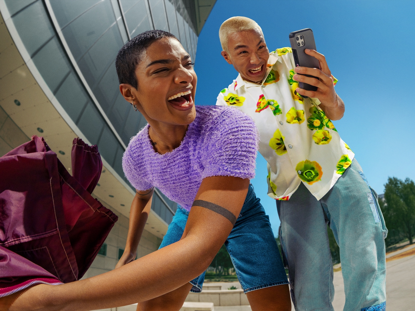 A woman and a man are outside, smiling as they look at a cell phone.