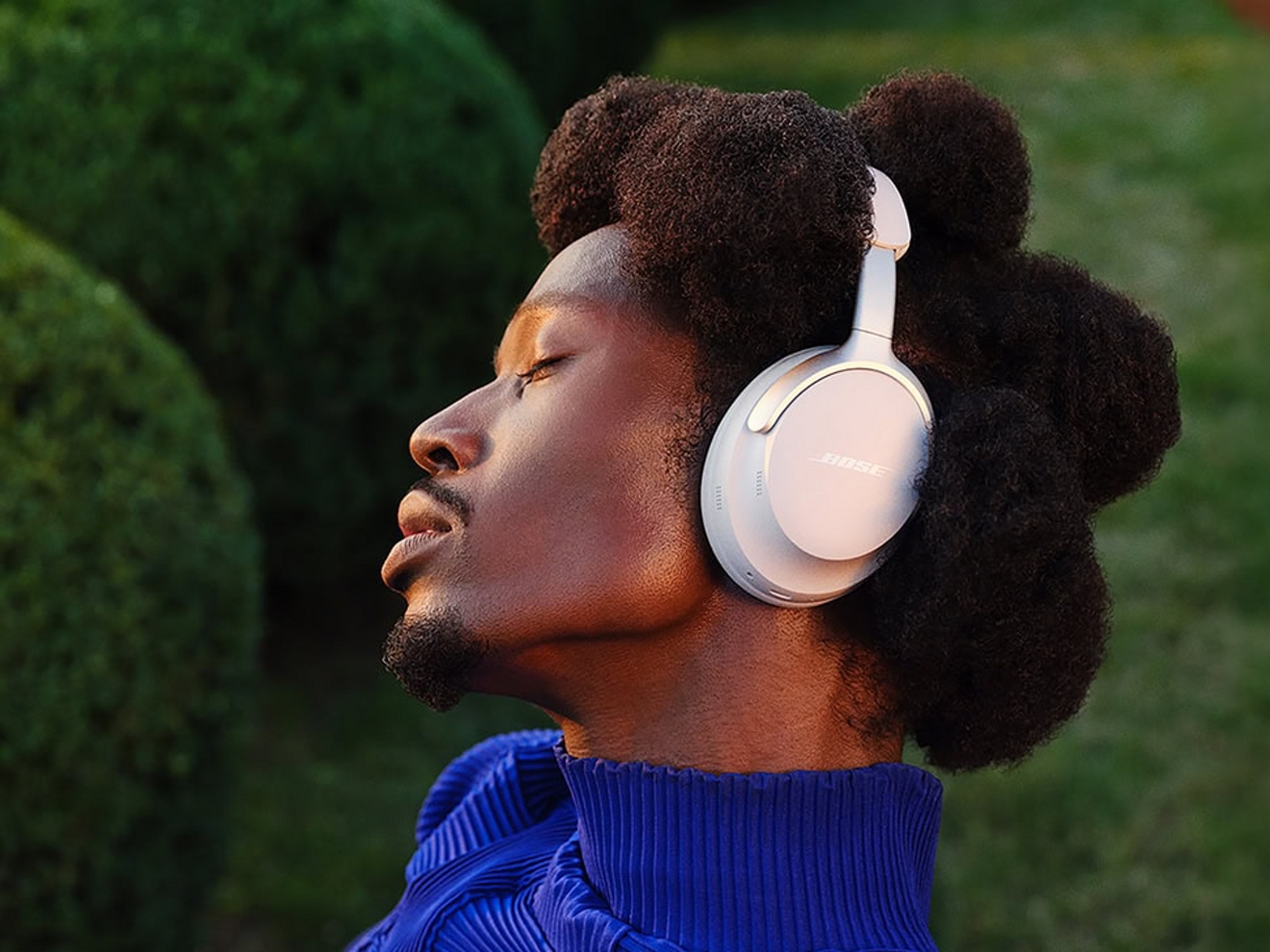 Bose QuietComfort wireless over-ear headphones in white, worn outdoors against a backdrop of green bushes.
